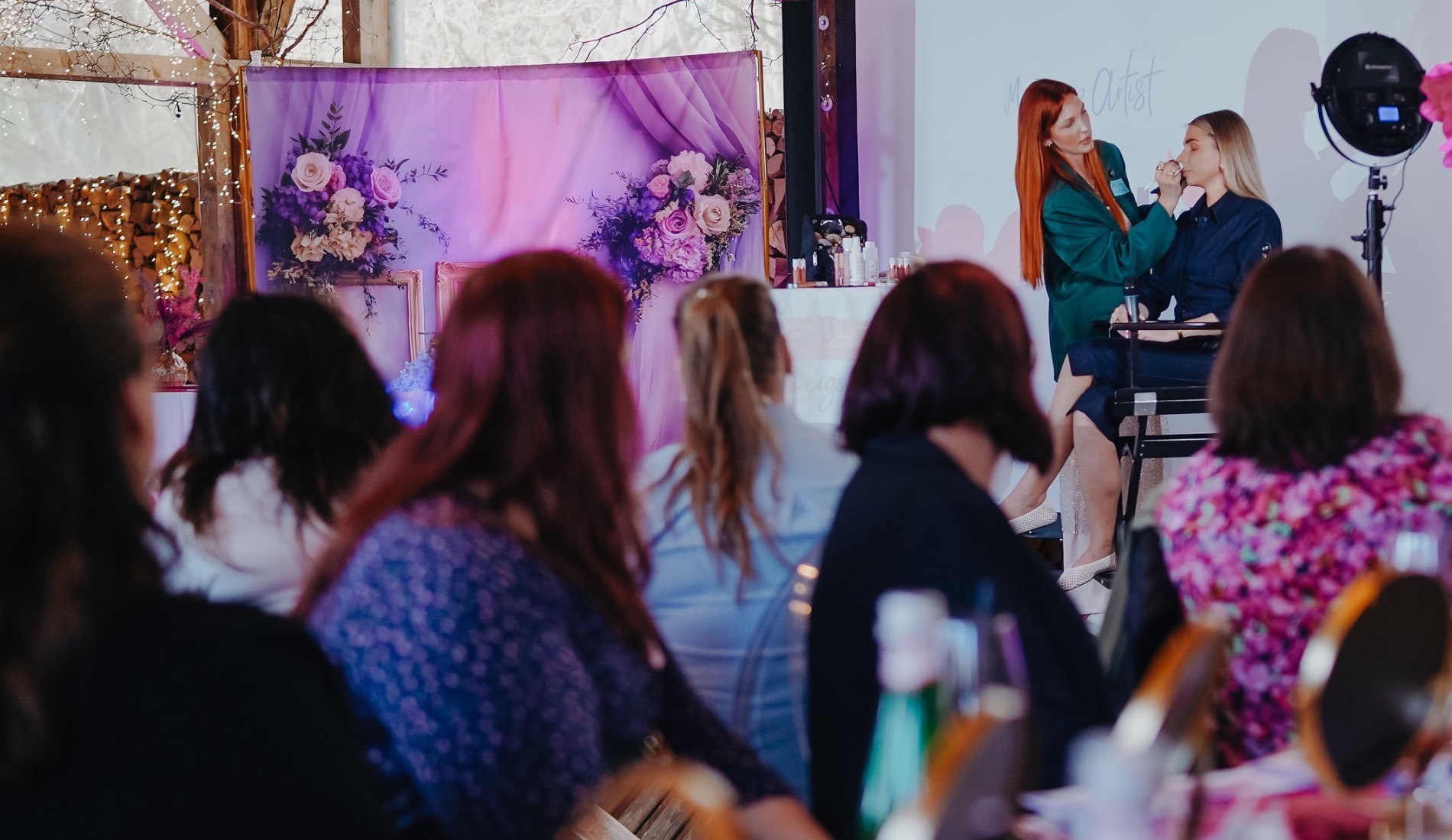 Make-up-Demonstration vor Publikum bei einem KLUGE-BEAUTY-Event: zwei Frauen auf einer Bühne vor einer Leinwand, davor Gäste an einer langen Tafel mit Spiegeln und Produkten