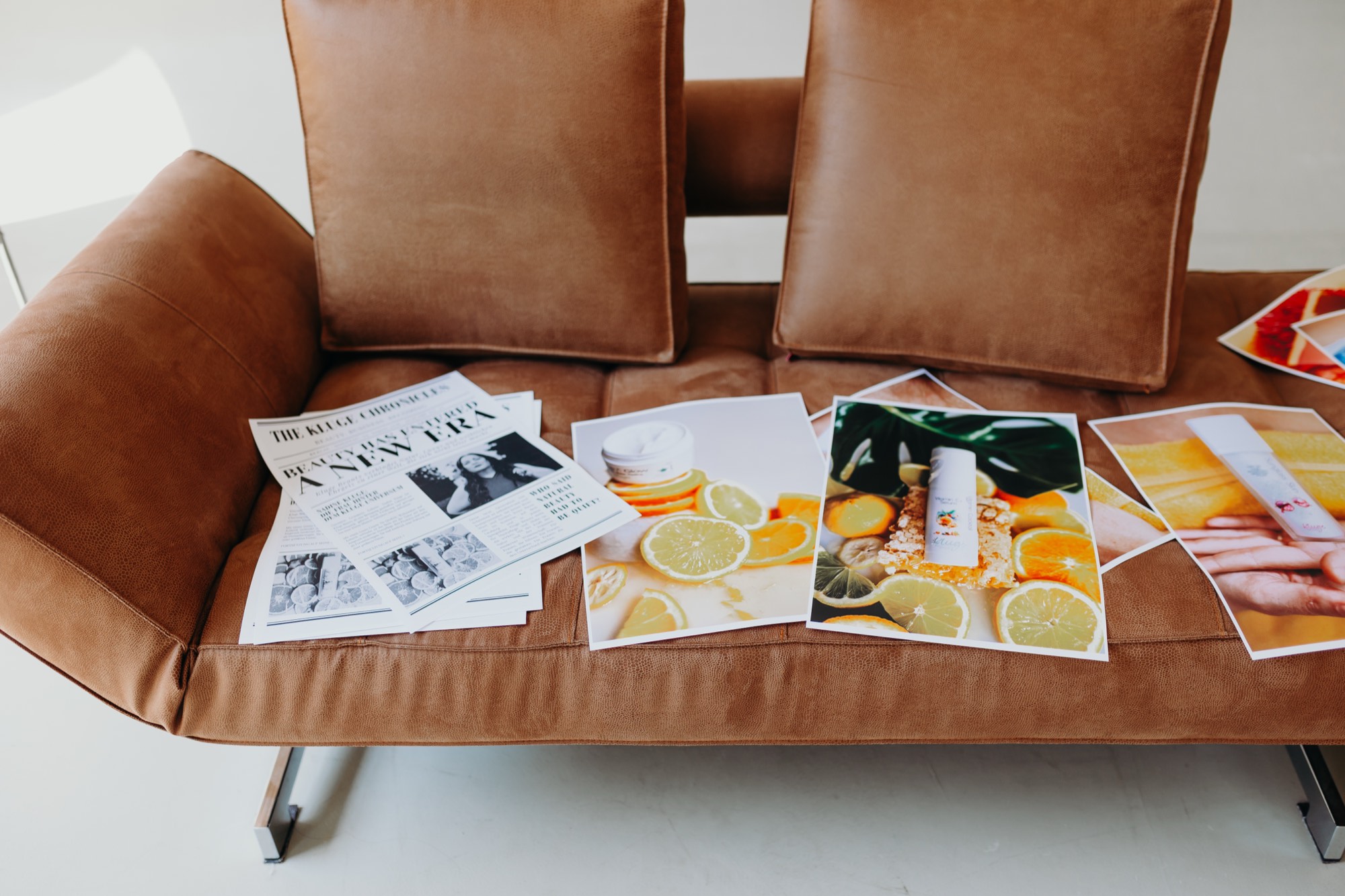 Zeitung „The Kluge Chronicles“ und großformatige Produktdrucke mit Zitronen und Orangen auf einem cognacfarbenen Ledersofa